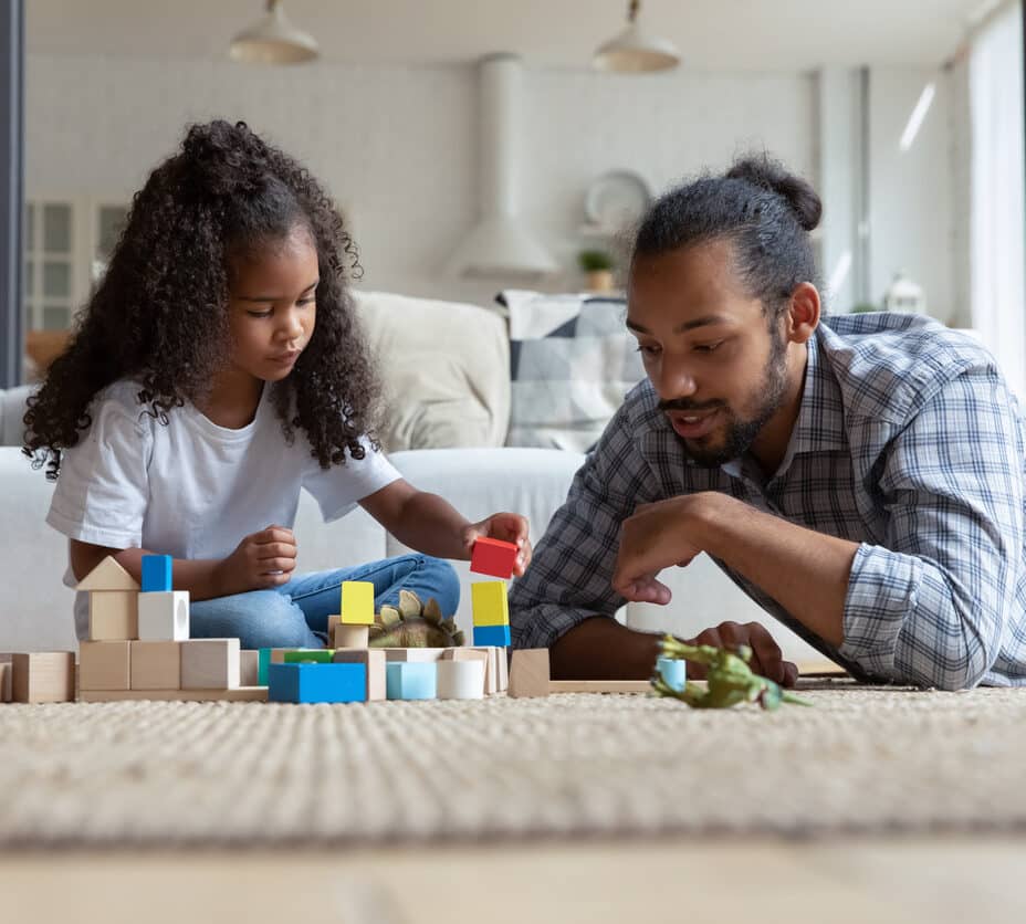 Interested kid building with wooden blocks with caring young father, sitting together on floor carpet in living room, enjoying playing at home.