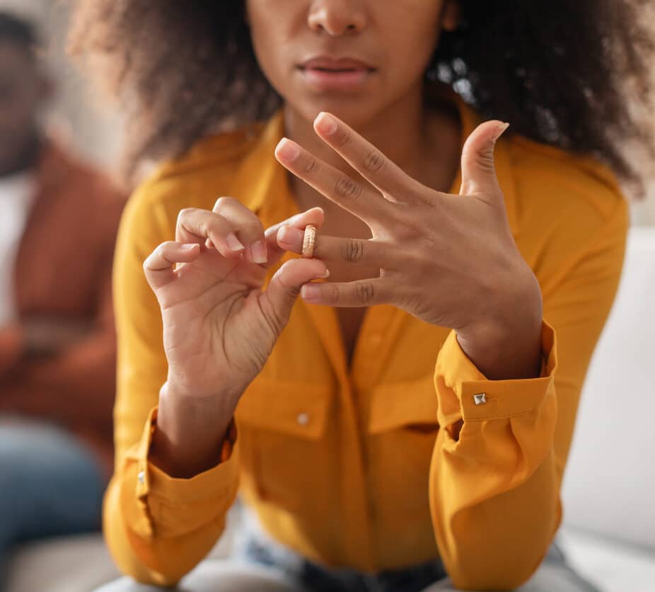 Divorce Concept. Unhappy black woman taking off wedding ring sitting next to husband indoors, cropped shot, selective focus. End of marriage or engagement.